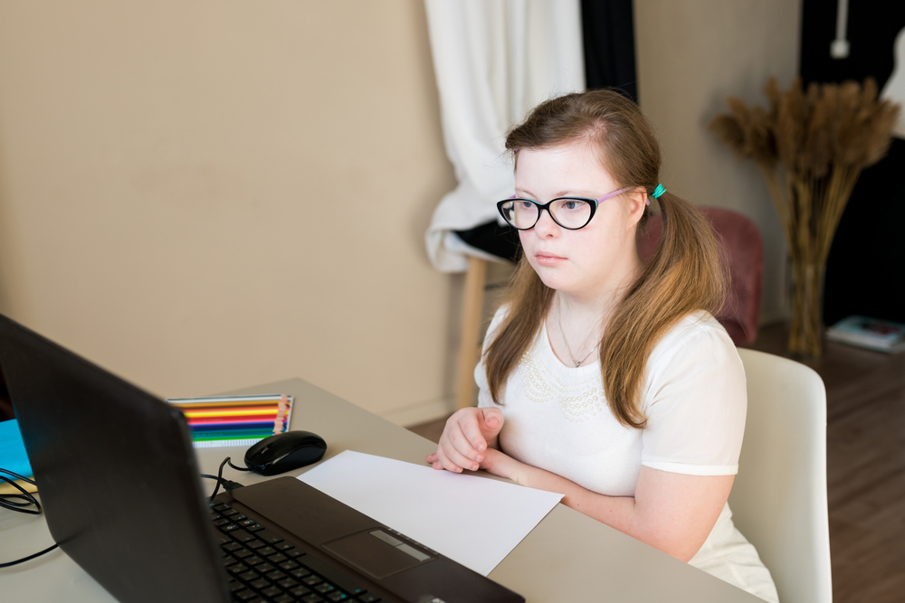 Teenage girl with Down syndrome wearing eyeglasses uses laptop to learn online at home.